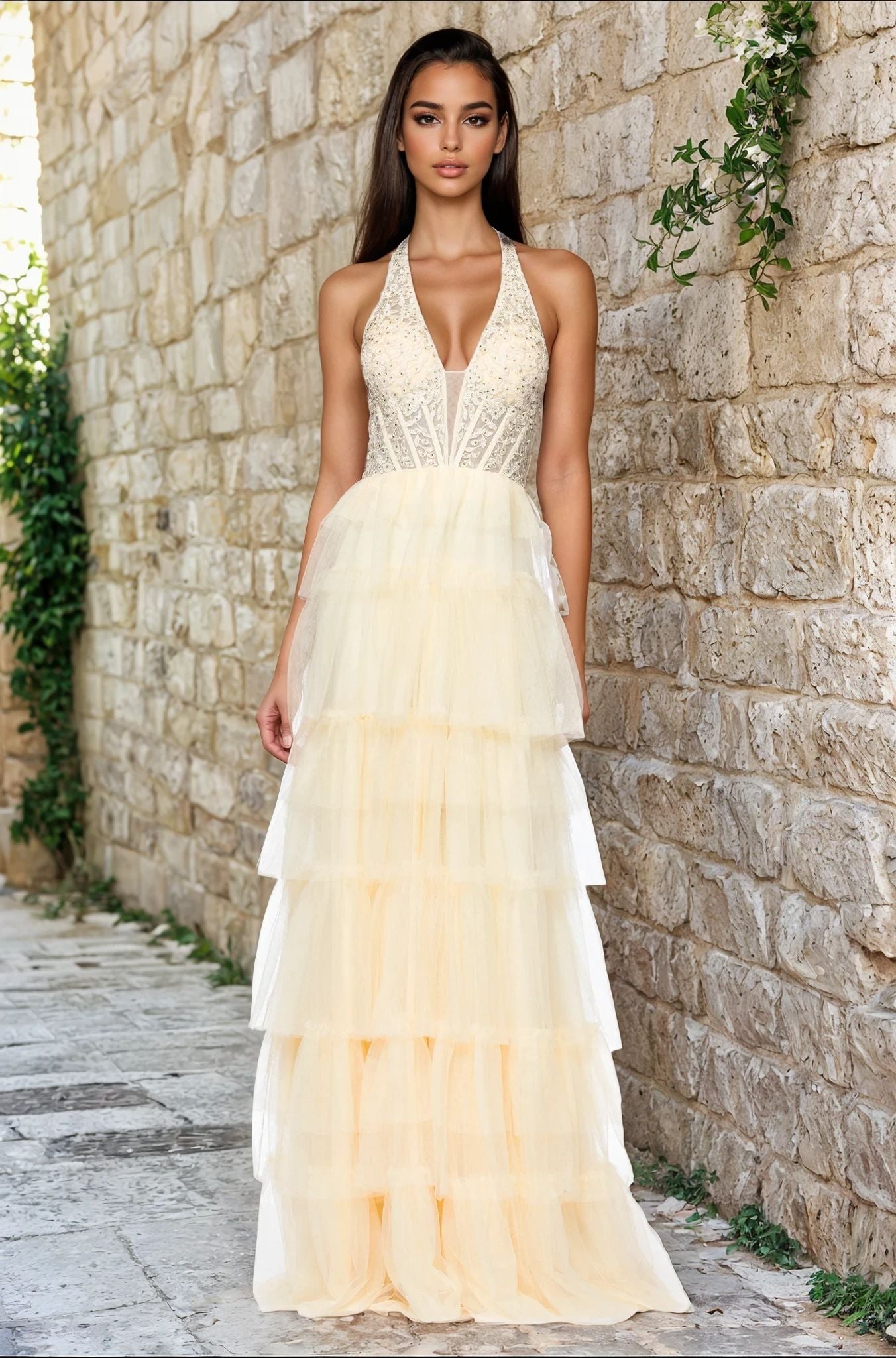 A woman stands outdoors against a stone wall in the Blondie Nites 5349BN long yellow halter prom gown, featuring a beaded bodice and layered pale yellow tulle skirt.